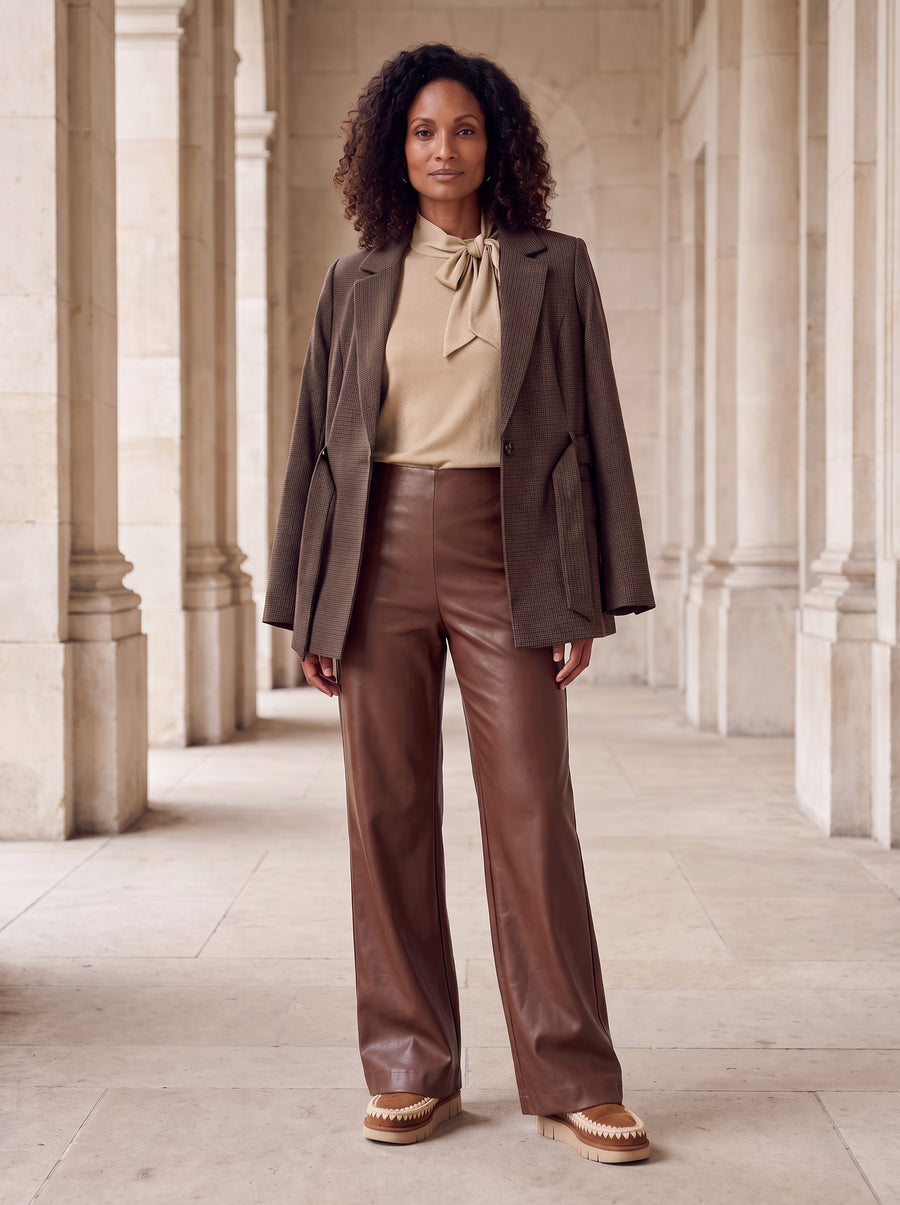 Model in bruine geruite Faya Blazer over de gouden Skyler Top met strik en cognac leren Olivia Pants met cognac suede Mou-laarsjes in een stenen zuilengang