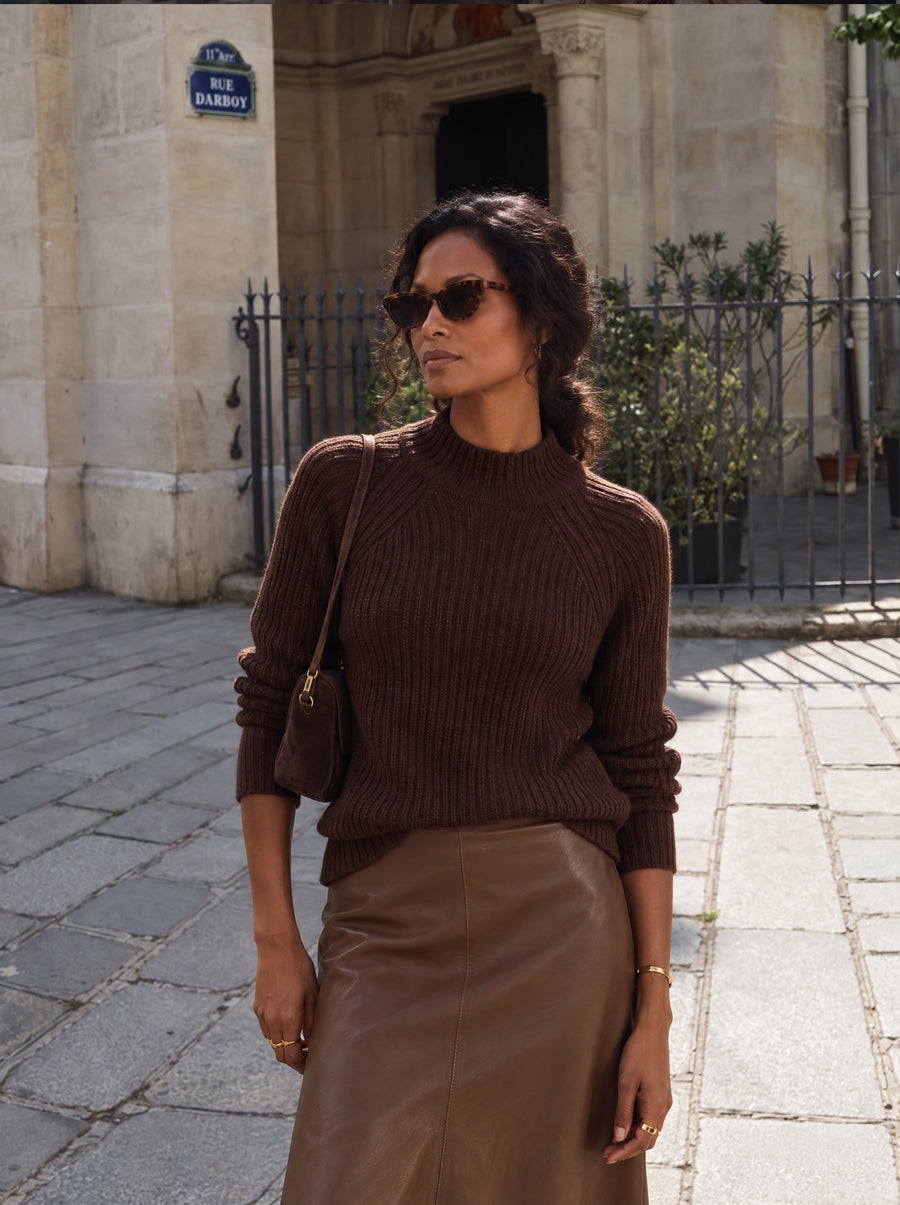 Vrouw draagt de Lize Pullover in bruin, een grof geribde trui met opstaande kraag, op de Sky Skirt in cognac van vegan leather